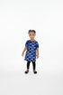 A young child standing barefoot on a shag rug in a living room, wearing a blue and white geometric patterned dress with orange trim and black leggings.