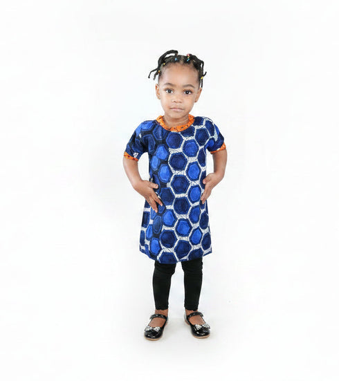 A young child standing in a living room wearing a blue and white patterned dress with orange trim, black leggings, and braids.