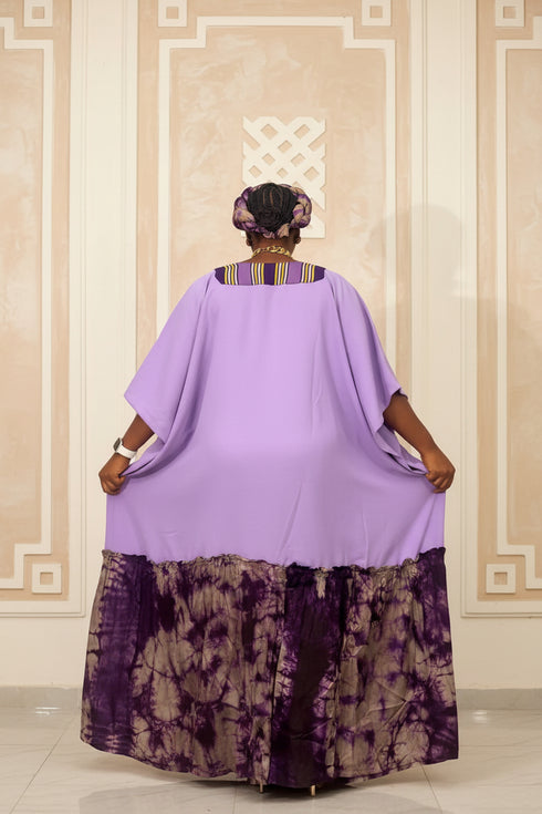 A person shown from the back wearing a loose lavender-purple kaftan-style dress with a patterned purple and black skirt, standing indoors in front of dark horizontal blinds. Large overlaid text reads, 'Which color is your fav? Price - N50,000 each WhatsApp 2347035008482'.