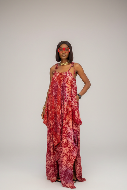 A woman models a red and orange batik-patterned maxi dress with spaghetti straps in a runway show setting, with two other models in matching dresses blurred in the background.