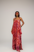 A woman models a red and orange batik-patterned maxi dress with spaghetti straps in a runway show setting, with two other models in matching dresses blurred in the background.