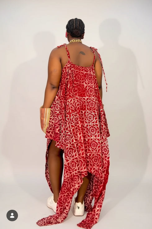 A woman viewed from behind wearing a red and pink patterned high-low dress with thin straps, white sneakers, and a gold chain necklace, standing in front of a black and white striped wall.