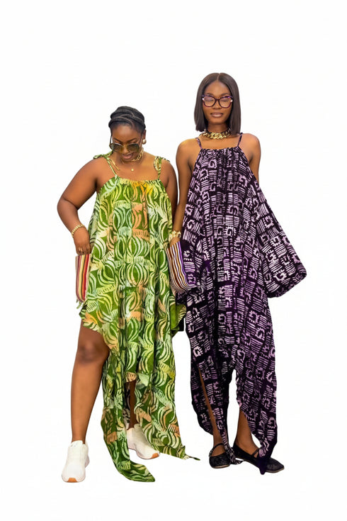 Two women modeling matching high-low dress sets with African wax print. The woman on the left wears a green and yellow patterned dress with white sneakers, and the woman on the right wears a purple and white patterned dress with black shoes.
