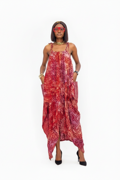 A woman modeling a sleeveless, high-low maxi dress with a red, pink, and orange abstract batik pattern, set against a plain white background.