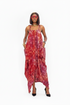 A woman modeling a sleeveless, high-low maxi dress with a red, pink, and orange abstract batik pattern, set against a plain white background.