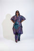 A woman modeling a matching two-piece batik tunic and trouser set in a home setting. The set features a vibrant multicolor pattern with purple, teal, pink, and black.