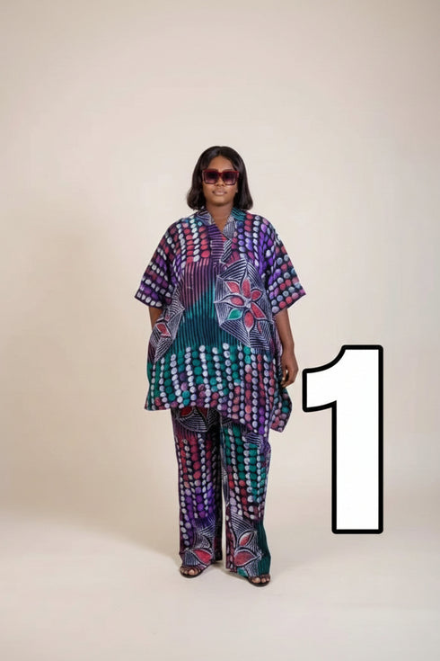 A woman modeling a matching two-piece outfit with a vibrant, multicolored African wax print featuring purple, green, blue, and red patterns, set against a plain beige studio background.