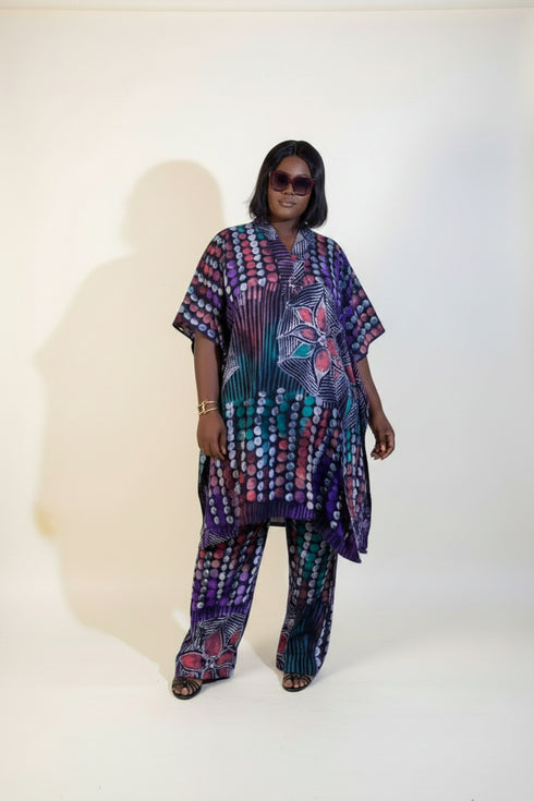A woman modeling a matching two-piece printed outfit with a loose kaftan-style top and wide-leg pants, standing indoors against a beige wall with framed wall art and a plant. A phone screen overlay appears in the lower left showing a smaller preview of the outfit.