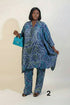 A woman modeling a blue and white patterned matching tunic and trouser set in an indoor setting with a framed wall collage, a potted plant, and a green ottoman. She is holding a bright blue handbag.