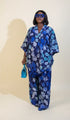 A woman wearing a blue and white floral pajama set stands in a room with framed wall art, a plant, and a green chair. A phone screen overlay in the bottom left shows a smaller version of the pajama set.