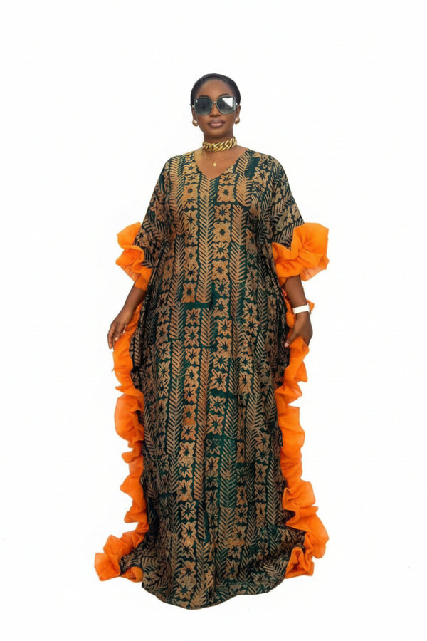A woman modeling a long, loose-fitting patterned dress with bright orange ruffled sleeve and side details, standing in front of a black-and-white striped wall. The dress has a dark green and beige geometric print.