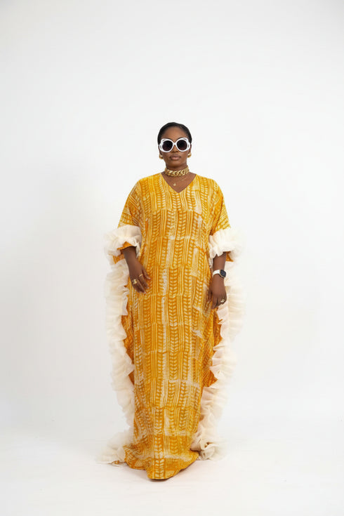 A person wearing a bright yellow and orange patterned traditional dress with white ruffled trim, standing in front of a black slatted wall. The image appears to be a social media post with text overlays showing pricing and WhatsApp contact information.