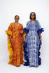 Two women modeling traditional African dress garments in a social media post. The woman on the left wears a vibrant orange and yellow patterned dress, and the woman on the right wears a blue and white patterned dress with dark blue ruffled trim.