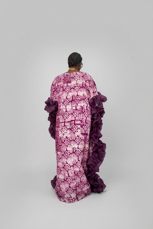 A woman shown from behind wearing a long magenta and purple patterned dress with ruffled sleeves, standing indoors in front of a dark paneled wall. A phone screen inset appears at the bottom left, and text overlays advertise the dress price and WhatsApp contact.