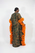 A woman wearing a long, loose caftan dress with a green and black geometric pattern and bright orange ruffled trim, shown from the back in an indoor setting with a striped wall behind her.