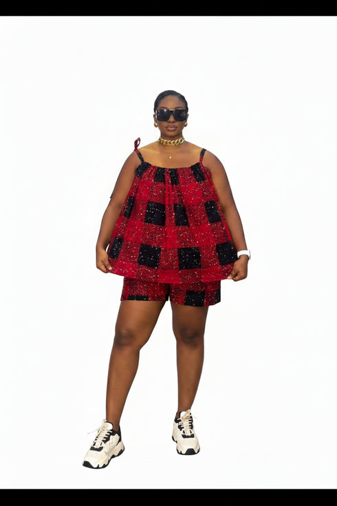 A woman modeling a red and black checkered sequined romper set, consisting of a sleeveless top and matching shorts, paired with white sneakers. She is standing in front of a white wall with black horizontal panels.