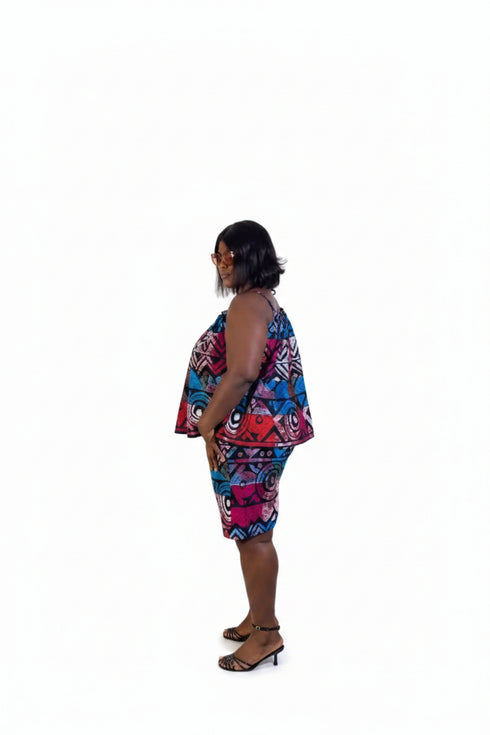 A woman stands in profile wearing a colorful sleeveless dress with a blue, red, and pink pattern, set against a beige wall with framed art and a potted plant.