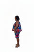 A woman stands in profile wearing a colorful sleeveless dress with a blue, red, and pink pattern, set against a beige wall with framed art and a potted plant.