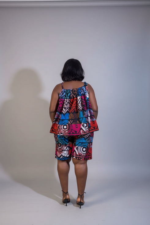 A woman stands with her back to the camera wearing a colorful sleeveless top and matching patterned shorts set. The room has a beige wall with framed wall art, a green plant, and a green ottoman.