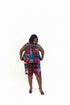 A woman modeling a matching two-piece sleeveless top and shorts set with a vibrant multicolor floral print, standing in an indoor room with framed wall art and a plant.