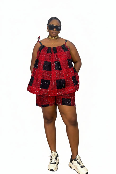 A woman modeling a red and black checkered sequined two-piece set consisting of a sleeveless top and matching shorts, paired with white sneakers and sunglasses, standing in front of a white wall with black horizontal blinds.