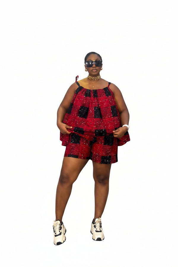 A woman modeling a red and black checkered sequined sleeveless top and matching shorts set, standing in front of a gray and white striped wall. She is wearing white and black sneakers, gold jewelry, and sunglasses.