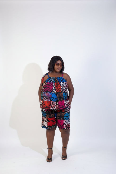 A woman modeling a colorful sleeveless floral romper in front of a beige wall with framed wall art and a green plant.