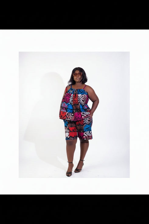 A woman modeling a colorful sleeveless two-piece matching set featuring a vibrant abstract pattern in red, blue, pink, and yellow, standing in an indoor setting with a beige wall, framed prints, and a potted plant.