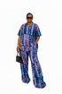 A woman modeling a matching short-sleeve button-front top and wide-leg trousers set in a blue, purple, and teal geometric pattern, styled with sunglasses, a gold choker, a gold bracelet, and a small black handbag against a plain white background.