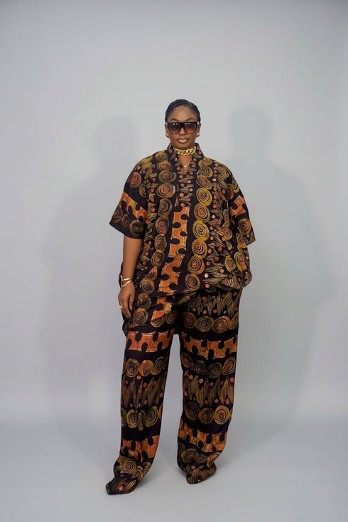 A person modeling a matching short-sleeve button-up shirt and wide-leg pants set with a bold circular and geometric pattern in orange, brown, and black, photographed against a plain light gray studio background.