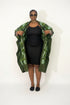A woman modeling a green patterned open-front jacket or coat over a black dress, standing against a plain white studio background.