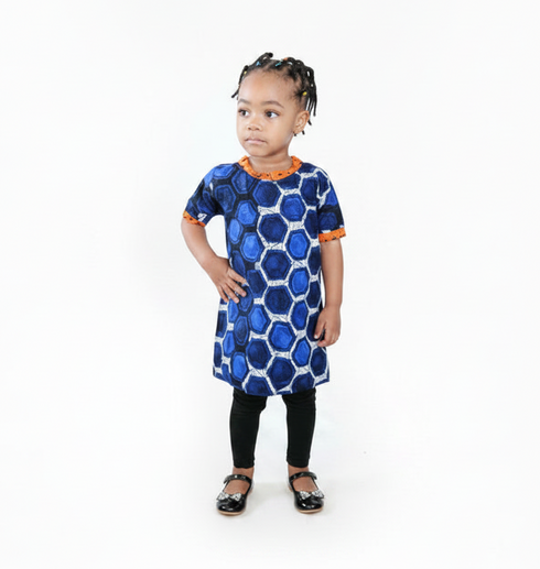A young child modeling a short-sleeve blue and navy patterned tunic dress with orange trim, paired with black leggings and sandals, standing against a seamless white background.