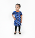 A young child modeling a short-sleeve blue and navy patterned tunic dress with orange trim, paired with black leggings and sandals, standing against a seamless white background.