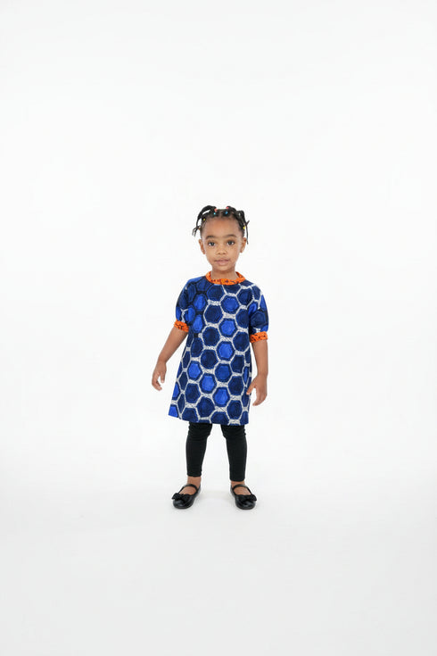 A young child standing barefoot on a shag rug in a living room, wearing a blue and white geometric patterned dress with orange trim and black leggings.