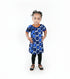 A young child standing in a living room wearing a blue and white patterned dress with orange trim, black leggings, and braids.