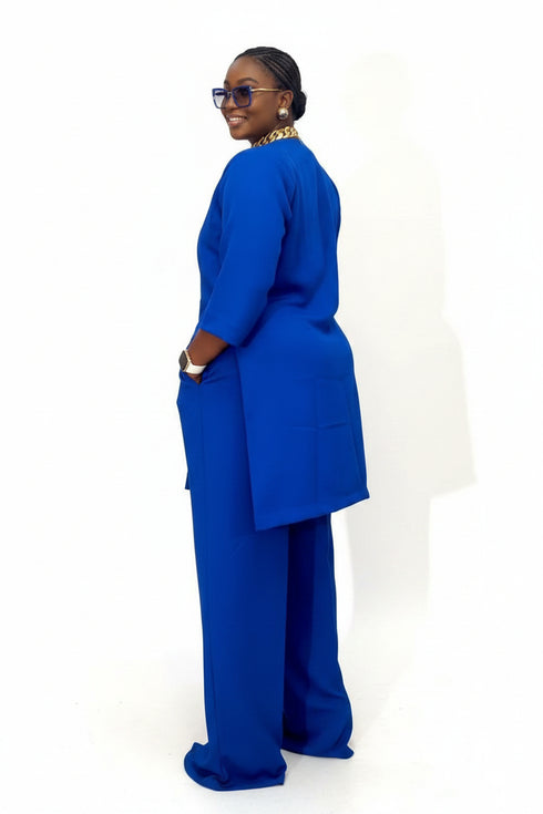 A woman wearing a bright royal blue two-piece outfit with a long-sleeve tunic and matching wide-leg pants, standing in front of a black-and-white striped wall. Social media interface overlays are visible on the right and bottom.