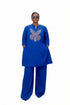 A woman modeling a royal blue two-piece outfit consisting of a long-sleeve tunic with a glittery butterfly design on the chest and matching wide-leg trousers, standing in front of a black and white striped wall.