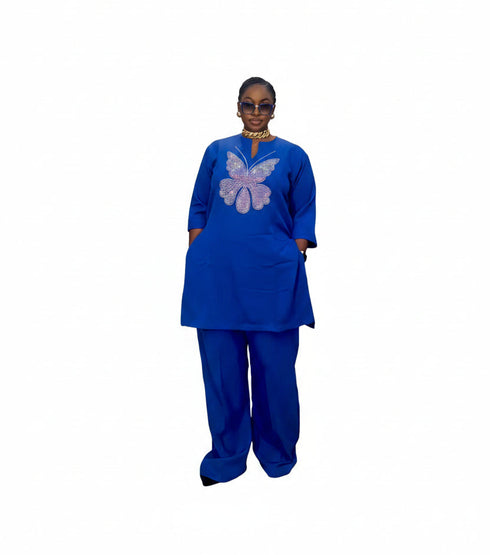 A person wearing a royal blue traditional outfit with a purple butterfly embroidery on the chest, standing in front of a black slatted wall. The image is a mobile screenshot with text overlays showing a price and WhatsApp contact, along with social media interaction icons.