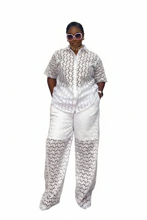 A woman modeling a matching white two-piece set with a black geometric pattern, consisting of a short-sleeve button-up shirt and wide-leg trousers, standing in front of a black and white striped wall.