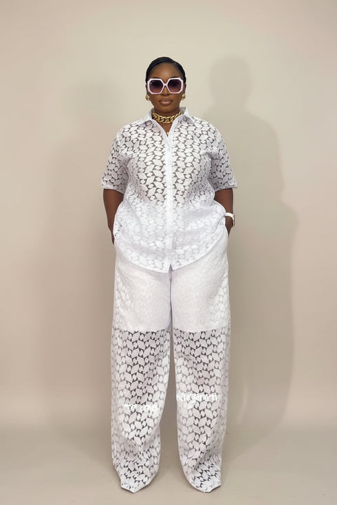 A model stands against a plain beige studio backdrop wearing a white short-sleeve button-up shirt and matching wide-leg pants with a black geometric lace-like pattern throughout.