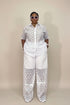 A model stands against a plain beige studio backdrop wearing a white short-sleeve button-up shirt and matching wide-leg pants with a black geometric lace-like pattern throughout.