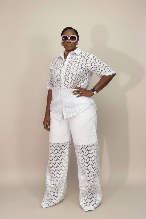 A model wearing a white two-piece matching set with a floral lace pattern, featuring a short-sleeve button-up shirt and wide-leg pants, posed against a solid beige studio background.