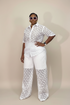 A model wearing a white two-piece matching set with a floral lace pattern, featuring a short-sleeve button-up shirt and wide-leg pants, posed against a solid beige studio background.