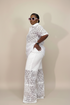 A woman modeling a white two-piece outfit consisting of a short-sleeve button-front lace top and matching wide-leg lace pants, photographed against a plain beige studio background.