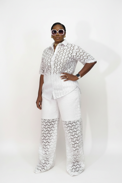 A model wearing a white two-piece outfit with a short-sleeve button-up shirt and matching wide-leg pants, both featuring a textured crochet lace pattern. She is posing against a plain white studio background.