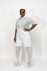 A model wearing a white two-piece outfit with a short-sleeve button-up shirt and matching wide-leg pants, both featuring a textured crochet lace pattern. She is posing against a plain white studio background.