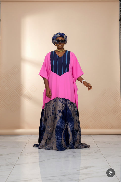 A woman modeling a vibrant pink and navy blue traditional dress with a tie-dye patterned skirt, standing against a black and white striped wall.