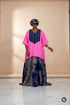 A woman modeling a vibrant pink and navy blue traditional dress with a tie-dye patterned skirt, standing against a black and white striped wall.