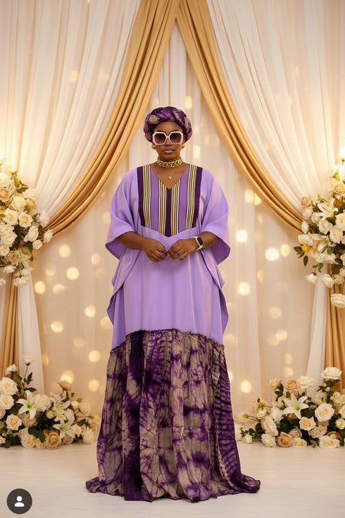 A woman wearing a lavender purple traditional outfit with a matching headwrap, white sunglasses, and a patterned purple and beige skirt, standing in front of a black and white striped wall.