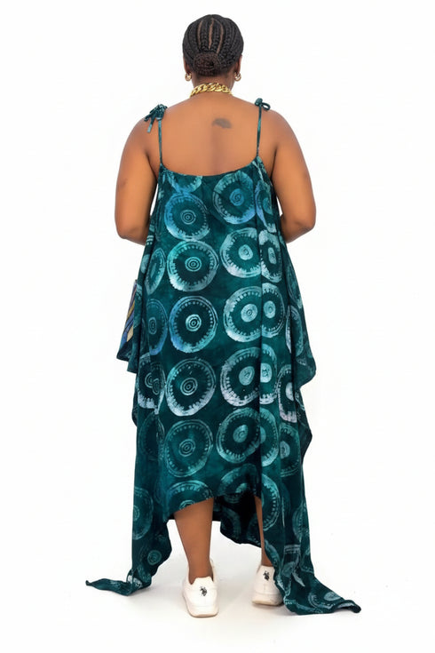 A woman viewed from behind wearing a sleeveless high-low dress with a teal and dark green circular pattern, standing in front of a gray and white striped wall.